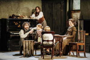 Production photo by Manuel Harlan. Nancy Carol sits at the table with two children a girl and a boy. Rory Kinnear is crouched down with his arm on the back of one of the wooden chairs, looking at the children. Oliver Chris is on the background with his elbow leaning on an old brown wooden piano.