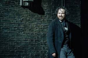 Production photo by Manuel Harlan. A dark brick wall with Rory Kinnear leaning against it. His hair is curly and hanging in his face and he has a beard. He appears to be smiling but looks wild.