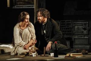 Production photo by Manuel Harlan. Laura Elphinstone, in a baggy off white long night gown, and Rory Kinnear are sat on the floor. Rory is looking at Laura, she is looking into the distance. There's a bottle of win on the floor next to them.