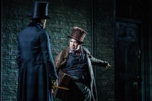 Production photo by Manuel Harlan. Nicholas Burns has his back to the camera and Milton Yerolemou is facing him, arm outstretched and looking angry. Both wear top hats and overcoats in black and brown.