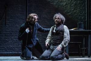 Production photo by Manuel Harlan. Oliver Chris kneels next to Rory Kinnear with his arm on his shoulder. Oliver wears and over coat and is looking at Rory. Rory is wearing a shirt and old sweater vest and looks exhausted on the floor.