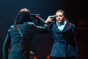 Production photo of Julius Caesar. Taken from behind Michelle Fairley we see her and Adjoa Andoh, both in black coats, both with their right hand holding a gun to their own heads.
