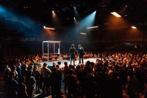Production photo of Julius Caesar. A wide shot of the auditorium. In a raised platform amongst one hundred standing audience members Michelle Fairley and Adjoa Andoh shake hands.