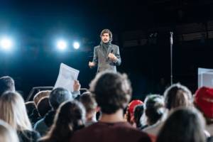Production photo from Julius Caesar. Taken from the audience we see the back of peoples heads, someone is holding up a poster that we can't see. On a raised platform stands Ben Whishaw in a grey smart coat and stripey scarf holding a microphone like he's making a speech. He looks worried.