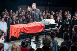 Production photo from Julius Caesar. On a slightly raised platform in the middle of crowds of audiences. David Morrissey stands on the platform above an open casket with David Calder lying dead as Caesar. Morrissey is holding his right hand up which is covered in blood.