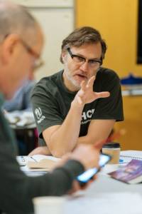 Rehearsal photo. David Morrissey is sat at and leaning on a table. One of his hands is raised and open as if explaining something. He's wearing a graphic t-short and glasses.