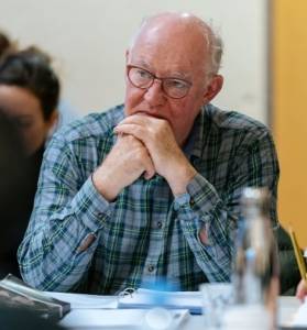 Rehearsal photo. David Calder in a blue chequered shirt and glasses, he's sat at a table with his elbows resting on its surface and his hands together covering his mouth.