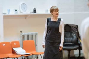 Rehearsal photo for Nightfall. Clare Skinner stands in a white top and blue long smock type dress, she looks angry. In the background of the room are 3 plastic orange chair and a leather computer chair.