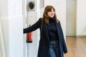 Rehearsal picture for Nightfall. Ophelia Lovibond is leaning against a wall smiling. She's in blue jeans, a black turtle neck and black smart coat.