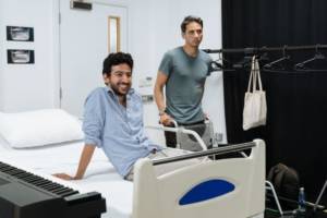 In the rehearsal room Manish Gandhi sits on a hospital bed leaning back on one arm, he's laughing. To his side is Gary Wood who is stood behind a wheelchair holding the handles. There's a keyboard to the left of the photo and a clothes rail to the right with empty hangers.