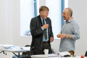 Alan Bennett in a navy suit stands talking to Director Nicholas Hytner who's in a blue and white stripey shirt. Both are side on and between two trestle tables covered in scripts, papers and coffee cups.
