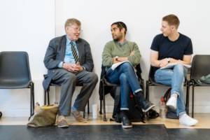Against a plain white wall, Alan Bennett, Sacha Dhawan and Samuel Barnett sit on plastic chairs. Beneath them are their bags, coffee cups and water bottles. Alan Bennett is in a navy suit with his hands together looking at the other two as if telling a story, they're looking back and listening.
