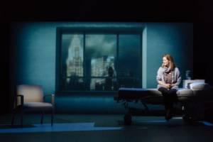 Production photograph from My Name is Lucy Barton. Laura Linney is sat on a hospital bed looking to the side at an empty hospital chair. Behind the scene is large hospital window frames with a projection of the city through it, you can see the Empire State Building.