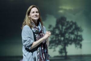 Production photograph from My Name is Lucy Barton. She is stood with her hands held together up at her chest, she's looking off into the distance wistfully. She's wearing a grey long cardigan and purple light scarf is wrapped around her neck. The silhouette of a tree is projected behind her.