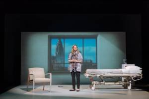 Production photograph from My Name is Lucy Barton. She stands centre stage with her hands together. There's a hospital bed to the right and a white hospital chair to the left. Behind is the large window frame filled with a projection of the view of New York, with the Empire State Building.