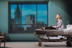 Production photograph from My Name is Lucy Barton. She is sat on the hospital bed with her legs out straight down the length of it. She looks out towards the camera. Behind is a large window frame filled with the projection of the view of New York.