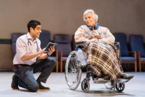 A low shot, in the blurred background are hospital chairs lined against the back wall. In the middle of the stage is Simon Williams in a wheelchair with a tartan blanket over his legs, he has headphones round his neck. He's looking at Sacha Dhawan who is on his knee next to him and reading a book to him.