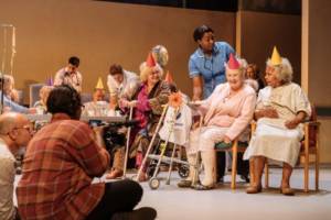 A low angle shot of the stage. The care home residents are sat in various formations, on plastic chairs or larger cushioned ones. They're all wearing colourful pointy party hats in reds, yellows and oranges. One woman has a zimmer frame with a sunflower attached to it. In the foreground out of focus is Nadine Higgin facing the scene holding up the large television camera.