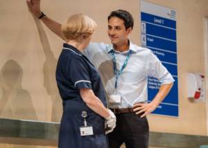 Close shot against the back wall of the set with the blue hospital signage. We see the back of Deborah Findlay in her blue nurses smock. With his arm outstretched leaning on the wall is Gary Wood in a shirt and suit trousers, he has a hospital lanyard on. He's smiling cheekily at Deborah.