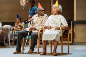 In the background is the remains of the party, there's a balloon with the number 80 on and an older lady eating cake. In the foreground Louis Mahoney (in a striped shirt and jeans but with a crutch in an orange party hat) and Cleo Sylvestre (in a hospital gown and a yellow party hat). Cleo is smiling and Louis is looking at her.