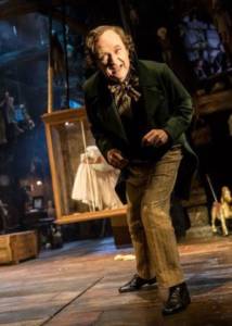 A close shot of the stage, in portrait angle. A full length Jim Broadbent stands leaning forward on one leg as if towards the audience, confiding in them. He's wearing pale brown subtly striped trousers, long smart black jacket and brown neckerchief. His hair is parted to one side and curled. The background looks like an attic and has wooden puppets hanging up around the place. Slightly obscured by the man we can see a glass and wooden box hanging from the ceiling with a girl leaning against the glass.