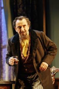 Close up of Phil Daniels as Charles Dickens. He's wearing period style clothes with striped trousers, a smart paisley waistcoat and brown long jacket. He's got a scraggly beard just on his chin and a moustache. In his right hand he's holding a small glass of whiskey. He looks angry with his eyebrows furrowed and teeth bared.