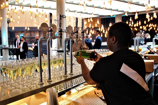 Picture from behind our bar. A person with a black t-shirt with a white Bridge Theatre beam on its back is pouring white wine. The bar has filled win glasses covering it. There are a few people chatting in the background and it is light.