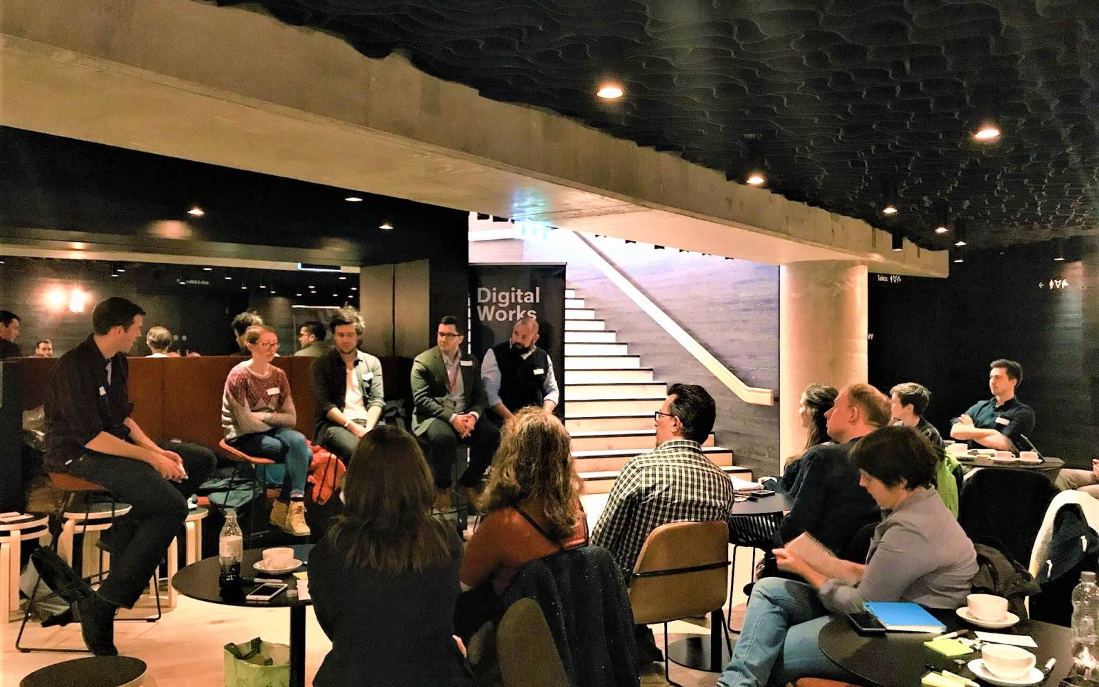 Picture of our lower foyer filled with people sat on high stools and lower chairs, there are tea cups and saucers on a round table and there is light coming in from the stairs up main foyer.