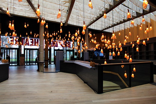 Picture of our main foyer from the back. Our copper wire lights are lit, hanging from the ceiling and natural light is also coming from above. Above our clear glass entrance doors the name Bridge Theatre is in read, but backwards and facing outwards.