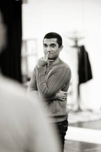 Black and white photo. In the foreground is shoulder that is blurry, then in focus further back is a man (Danny Ashok). He's got short dark hair and is wearing a cable knit jumper. His arms are crossed with one hand on this mouth.