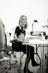 Black and white photo. Side on picture of a woman in a floral black shirt, and blonde shoulder length hair. She's sat at a table on a wheeled computer chair. She's holding a pen to her page and smiling.