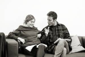Black and white photo. On a sofa sits a blonde woman (Leah Gayer) and a dark haired man (Sam Woolf). Both have their legs crossed and scripts on their laps. The woman is laughing with her head down and the man has his hand out, smiling, as if explaining something.