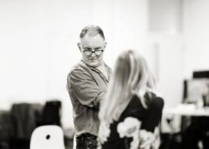 Black and white photo. In the blurred foreground is the back of Joanne Froggatt (black floral shirt and long blonde hair), the focus of the image is a man (Robert Glenister) who is wearing a shirt and black framed glasses, his arms are crossed and he's looking down over the top of his glasses.