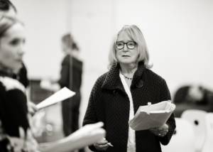 Black and white photo of a woman (Joanna David) wearing a chunky dark coat, and dark framed glasses. Her hair is shoulder length, blonde with a choppy fringe. She's holding a script in one hand and a pencil in the other. She looks despaired.