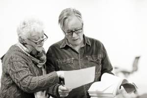 Black and white photo of a man (Jeff Rawle) holding scripts in both hands, and a woman (Sue Wallace) who's leaning over his shoulder pointing at one of the scripts. Sue is wearing a chunky knit cardigan, light scarf and dark framed glasses. Her hair is white, short and curly. Jeff is wearing a dark denim looking shirt and dark framed glasses.