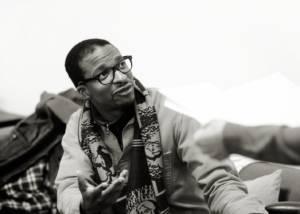 Black and white photo. Simon Manyonda is sat on a sofa and looking up at someone. Simon is wearing a patterned scarf and black framed glasses.