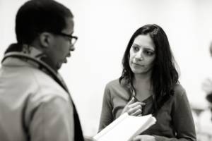 Black and white photo. In the foreground blurred is Simon Manyonda holding a script in his hands. The focus of the image is Vineeta Rishi who has long dark hair and is wearing a v neck jumper. She is looking at Simon concerned.