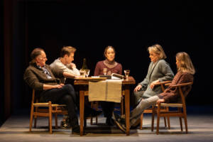 A rectangular table is on the sage with 5 people sat around it, the end closest to the camera has no one sat at it. Joanne Froggatt, with her blonde her back in a pony tail, is at the head of the table and everyone is looking to her. Everyone is in casual wear with knitted jumpers. There's a few wine glasses and a wine bottle on the table.
