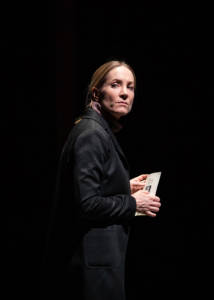 Portrait photo of Joanne Froggatt. The rest of the stage is obscured in darkness and there's a spotlight on her. She is wearing a smart black coat and her blonde hair is in a ponytail. She is looking back over her shoulder with her eyes squinted. She's holding the order of service in her hands but the details can't be seen.