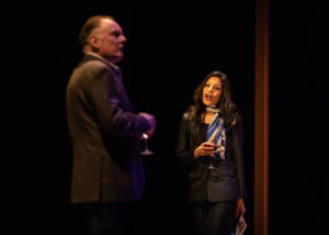 In the foreground of the photo is an out of focus man (Robert Glenister) who is looking outwards as if towards the audience. In focus further back in the photo is a woman with long dark curls (Vineeta Rishi). She's wearing a dark suit and a colorful scarf tied tight at the neck. She's holding a glass of wine.
