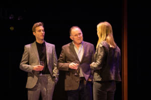 Two men are in focus here, Sam Woolf, young and tall with blonde short hair. He's wearing a grey suit with a causal black shirt underneath and is holding a glass of wine. Next to him is Robert Glenister, also holding a wine glass and wearing dark jeans, a white shirt and grey suit jacket. Blurred in the foreground is the back of Joanne Froggatt (blonde hair down and wearing dark skinny jeans and a leather jacket).