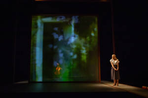 A wider shot of the stage. To the right is Joanne Froggatt who is wearing a mid length stripey skirt, white blouse and small heels. She's looking across the stage longingly with her hands together hanging in front of her. The stage has a partly transparent screen across it, on it is a projection of a green leafy print and on the other side you can just see a woman sat playing the cello.