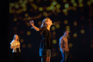The main focus of the photo is Leah Gayer who is centre stage. The photo captures her from the side, her arms are flung open, holding a glass in one. Her long blonde hair is behind her ears and she is wearing blue denim shorts and a smart black top that looks velvet. In the background we see a man and a woman who are out of focus and projected on the screen behind looks like fairy lights.