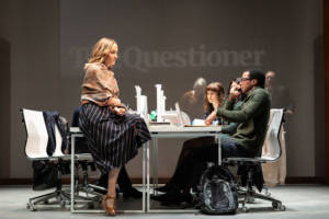 An office scene on the stage. On the screen at the back is projected the words The Questioner in a newspaper style font. Joanne Froggatt is sat on one of the desks in the striped skirt with a paisley patterned scarf wrapped around her. Simon Manyonda is sat at one of the desks on the phone and looking afraid.