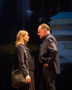 Portrait photo of Joanne Froggatt and Robert GlenisterJoanne Froggatt and Robert Glenister. They're both in smart blazers and Joanna has a leather bag on her shoulder. They're close and facing each other. Robert has his hands on his hips. On the screen behind you can just see a projection of a stack of books.