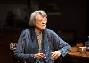 A close shot of a woman, Maggie Smith, sat at a round wooden kitchen table. She's got short grey hair and is wearing a long blue cardigan and blue scarf. She's looking to the side and holing a pair of glasses between her hands as if she's just taken them off and is closing them.