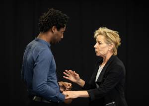 A side on picture of a man, Yoli Fuller, and a woman, Zoë Wanamaker, facing each other. Yoli is taller and Zoë reaches out her hands to him with a concerned look on her face. She's wearing the black blazer and he is in a collarless blue shirt.