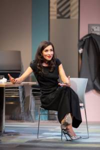 Zrinka Cvitešić is sat in a metal chair next to a long table. She's wearing a long smart black dress with snakeprint heels. Her legs are crossed and she is holding her hands and arms out as if asking a question with great emphasis.