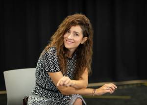 The background is a black curtain. Sat on a plastic chair is a woman, Zrinka Cvitešić, who has long curly brown hair. Her legs are crossed and she's leaning forward a bit resting on her legs. With her head titled to the side she's smiling.