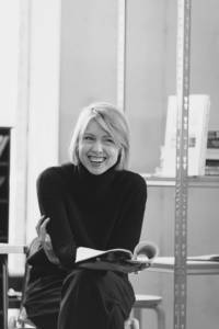 Black and white photo of a woman (Polly Findlay) in her 30's. She's wearing a black rollneck jumper and smart black trousers. Her legs are crossed and she's leaning on them, holding a script open in one of her hands. She has a blonde bob with a side parting and she's smiling as if she's laughing.